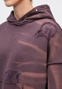 Camisola com capuz roxa, apresentando uma textura desgastada com detalhes desfiados e molas prateadas no capuz. Tecido macio e casual com um design descontraído.