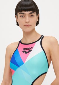 Woman with short dark hair wearing a multicolored high-neck swimsuit with black trim and an "arena" logo on the chest.