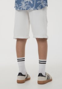 Pantaloni shorts in denim bianco con risvolti, abbinati a calzini bianchi con due strisce nere e sneakers beige con dettagli scuri.