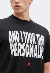 Νέος φοράει μαύρο T-shirt με μεγάλο λευκό κείμενο «AND I TOOK THAT PERSONALLY.» και μικρό κόκκινο λογότυπο Jumpman από κάτω.