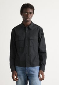 Mann iført en svart knapp-up skjorte med to brystlommer og lyseblå jeans, stående mot en enkel hvit bakgrunn.