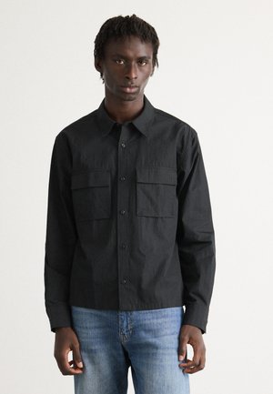 Man wearing a black button-up shirt with two chest pockets and light blue jeans, standing against a plain white background.