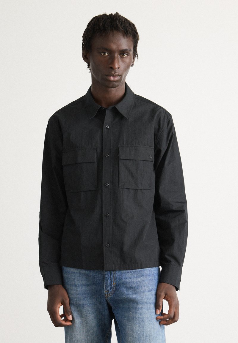 Uomo che indossa una camicia nera con bottoni, dotata di due tasche sul petto, e jeans azzurri chiari, in piedi contro uno sfondo bianco semplice.