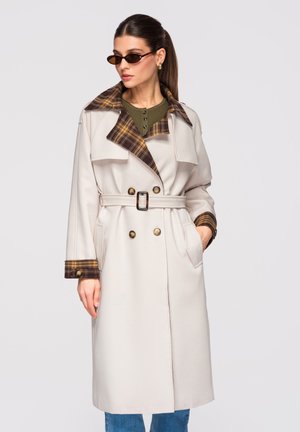 Femme portant un trench beige avec col et poignets à carreaux, taille cintrée, haut vert, jean bleu et petites lunettes de soleil foncées.