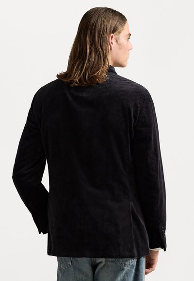 Jongere met schouderlang haar, gekleed in een zwarte corduroy blazer en een blauwe spijkerbroek, gezien vanachter tegen een witte achtergrond.