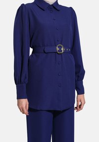 Blusa blu marino con colletto, maniche lunghe a sbuffo, chiusura con bottoni e una cintura coordinata con fibbia dorata, realizzata in tessuto testurizzato.