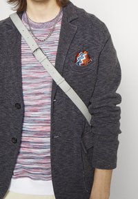Blazer grigio scuro con tessuto testurizzato, indossato sopra una camicia a righe multicolori. Il fazzoletto da taschino presenta motivi a zigzag in rosso e blu.