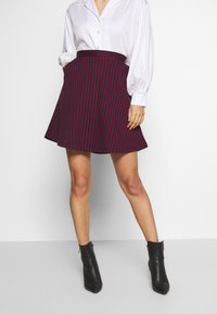 Vrouw in een witte blouse, marineblauwe rok met rode verticale strepen en zwarte enkellaarsjes, staand met één hand in de zak.