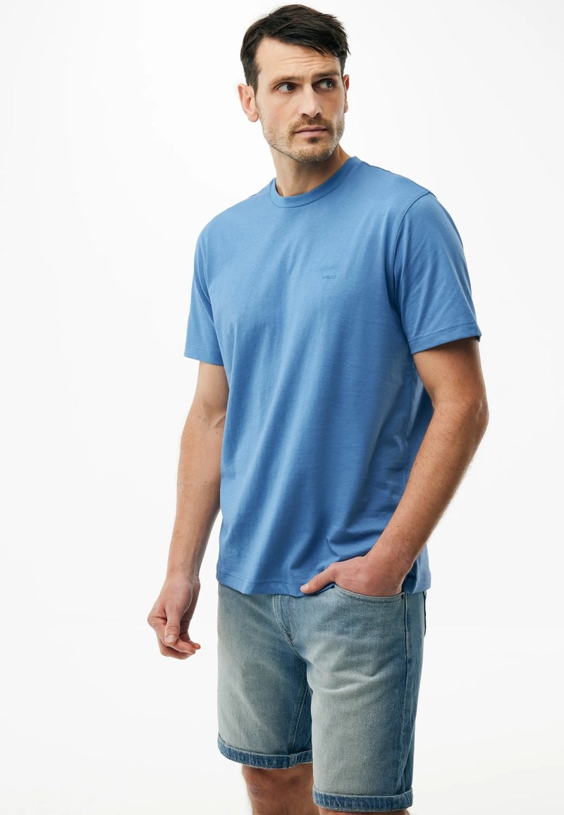 Hombre con camiseta azul de manga corta y pantalones cortos de mezclilla clara, de pie con una mano en el bolsillo y mirando hacia un lado.