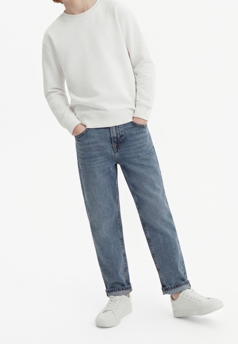 Personne portant une chemise blanche à manches longues, un jean bleu retroussé et des baskets blanches, debout les mains dans les poches.
