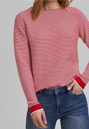 Kobieta ubrana w czerwono-biały, poziomo w paski top z długim rękawem, włożony do niebieskich dżinsów, z jedną ręką w kieszeni.
