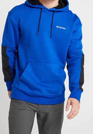 Hombre con una sudadera con capucha azul Columbia con parches negros en los codos y pantalones grises, de pie contra un fondo liso.