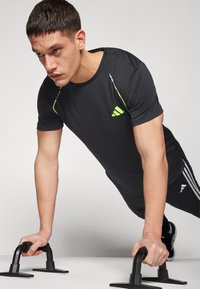 Sort atletisk t-shirt med neongrønne accenter og tre striber på bukserne. En person udfører armbøjninger ved hjælp af sorte armbøjningsstænger.