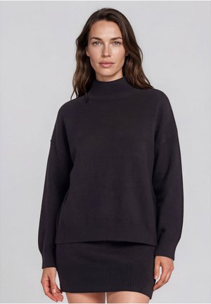 Femme aux longs cheveux bruns portant un pull noir à col roulé et une jupe en maille côtelée assortie, debout devant un fond gris clair.