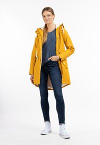 Parka galben impermeabil cu glugă căptușită cu fleece, detalii cu nasturi și șnur, purtată peste o cămașă cu dungi bleumarin și blugi negri. Tenisi albi.