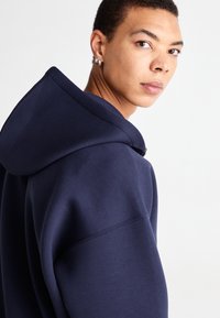 Le sweat à capuche bleu marine présente une coupe décontractée, une grande capuche et une texture douce. Les détails notables incluent des accents de couture et un design épuré et minimal.