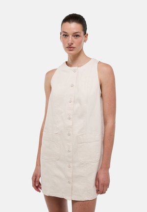 Femme vêtue d'une robe beige sans manches à boutons avec deux poches avant, debout devant un fond clair uni.