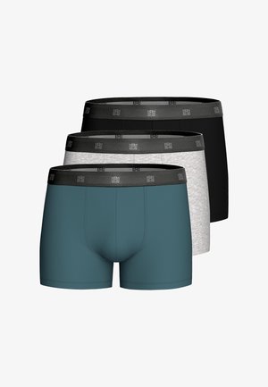 Gespierde man met kort haar die turquoise boxershorts met een zwarte tailleband draagt, staand tegen een effen lichtgrijze achtergrond.
