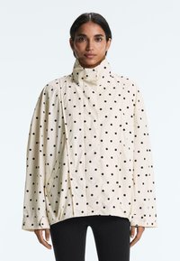Femme portant une veste blanche ample à col haut avec des pois noirs, associée à un pantalon noir, debout devant un fond blanc uni.
