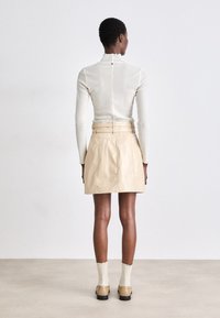 Haut crème à manches longues avec col volanté, assorti à une jupe en cuir plissée beige dotée d'une ceinture. Modèle vu de dos.