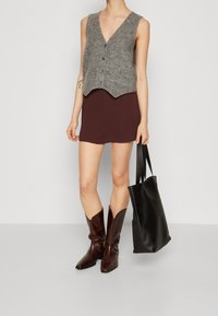 Szary dzianinowy kamizelka z dekoltem w kształcie litery V i guzikami z przodu, w zestawieniu z krótką brązową spódnicą. Brązowe kozaki do kolan i czarna torba na ramię dopełniają look.