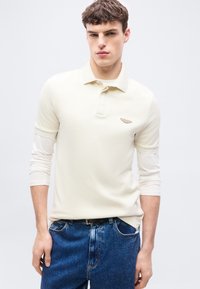 Νεαρός άνδρας που φοράει ένα μπλουζάκι πόλο μπεζ με κοντά μανίκια πάνω από ένα λευκό πουκάμισο με μακριά μανίκια και μπλε τζιν, στέκεται μπροστά σε ένα απλό ανοιχτόχρωμο φόντο.