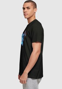 Camiseta negra con un gráfico de un personaje de dibujos animados azul; manga corta, cuello redondo, corte casual. Modelo visto de lado contra un fondo neutro.