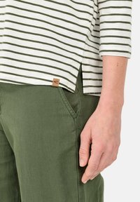 Gestreiftes, weißes und olivgrünes Hemd mit Dreiviertelärmeln, seitlichen Schlitzen und einem kleinen braunen Etikett. Kombiniert mit grünen, strukturierten Hosen.
