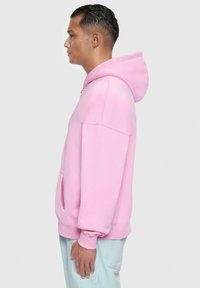 Light pink hoodie made of soft fabric; features a large hood, kangaroo pocket, and ribbed cuffs. Side view showcases loose fit and design.