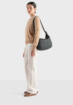 Bolso de tela gris para el hombro con una correa negra, que presenta una forma curva y un detalle sutil del logo. La modelo lleva un suéter beige y pantalones blancos.