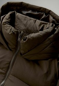 Veste isolante marron avec un col haut, fermeture éclair et tissu texturé. Présente un design rembourré et une doublure intérieure lisse.