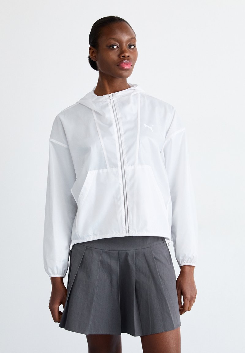 Veste coupe-vent blanche à capuche avec fermeture éclair, dotée d'une poche kangourou et de poignets élastiques, portée avec une jupe grise plissée.