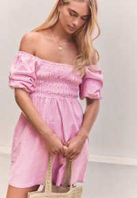 Vaaleanpunainen olkapäät paljastava mekko, jossa on liehukkeet hihansuissa, nyörityksellä muotoiltu yläosa ja pystysuora raidallinen kuvio. Malli pitelee punottua laukkua.