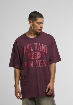 Jeune homme aux cheveux courts portant un T-shirt ample marron rouge avec le texte "KARL KANI 89 NEW YORK", debout devant un fond gris uni.