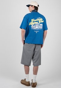 Jeune homme portant un t-shirt bleu à motif graphique, un short gris à fines rayures, une casquette beige, des chaussettes blanches et des chaussures marron, debout dos à la caméra.