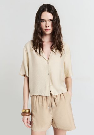 Chemise beige à manches courtes avec un devant boutonné, présentant un tissu texturé. Associée à un short beige à cordon de serrage. Bracelets en or et en bois portés.