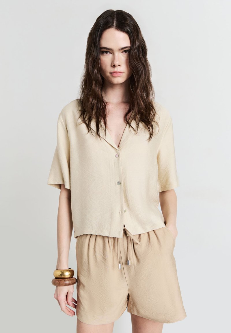 Camicia beige a maniche corte con apertura a bottoni, caratterizzata da un tessuto texturizzato. Abbinata a pantaloni corti beige con cordoncino. Bracciali in oro e legno indossati.