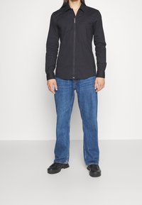 Zwart blouse met lange mouwen en een ritssluiting aan de voorkant, effen kleur en met kraag. Draagt met blauwe rechte jeans en zwarte schoenen.