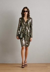 Robe enveloppante métallisée scintillante avec des manches longues, une taille nouée et une longueur à hauteur du genou. Associée à des sandales à talons hauts noires et des lunettes de soleil sombres.
