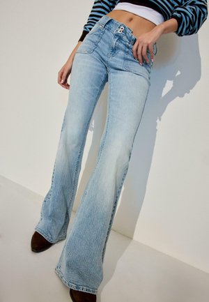 Person trägt hellblaue, hoch taillierte Schlagjeans und ein gestreiftes, kurz geschnittenes Langarmshirt, steht entspannt mit den Händen an einer hellen Wand.