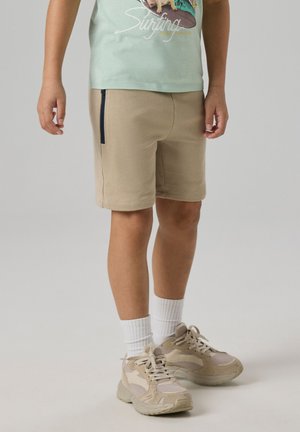 Enfant portant un short beige avec une poche zippée sur le côté, des chaussettes blanches et des baskets beiges, debout devant un fond uni.