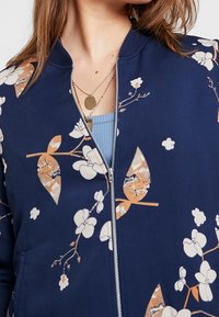 Námornícky bomber bunda s květinovými vzory v krémové a oranžové barvě. Zapínání na zip. Rybí límec. Lehký materiál s hladkou strukturou.