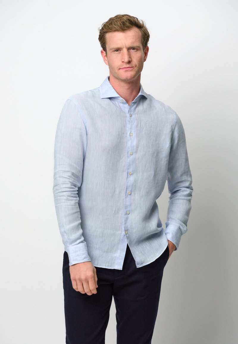 Camicia button-up in lino azzurro chiaro con maniche lunghe, caratterizzata da un colletto a punta e una texture sottile, abbinata a pantaloni scuri.