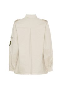 Beige bomullsskjorta med axelremmar, knäppta ärmslut och en veck bak. Har en broderad applikation på vänster ärm.