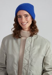 Beanie blu in maglia a coste; giacca bomber grigio chiaro con zip frontale e motivo trapuntato; maglione a collo alto crema sotto.