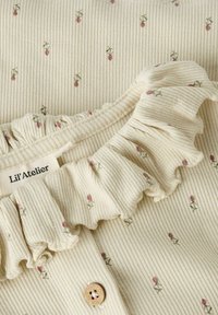 Crème-kleurige ribstof met kleine bloemenpatronen. Voorzien van een gegolfde halslijn en een houten knoopsluiting. Merkenlabel zichtbaar.