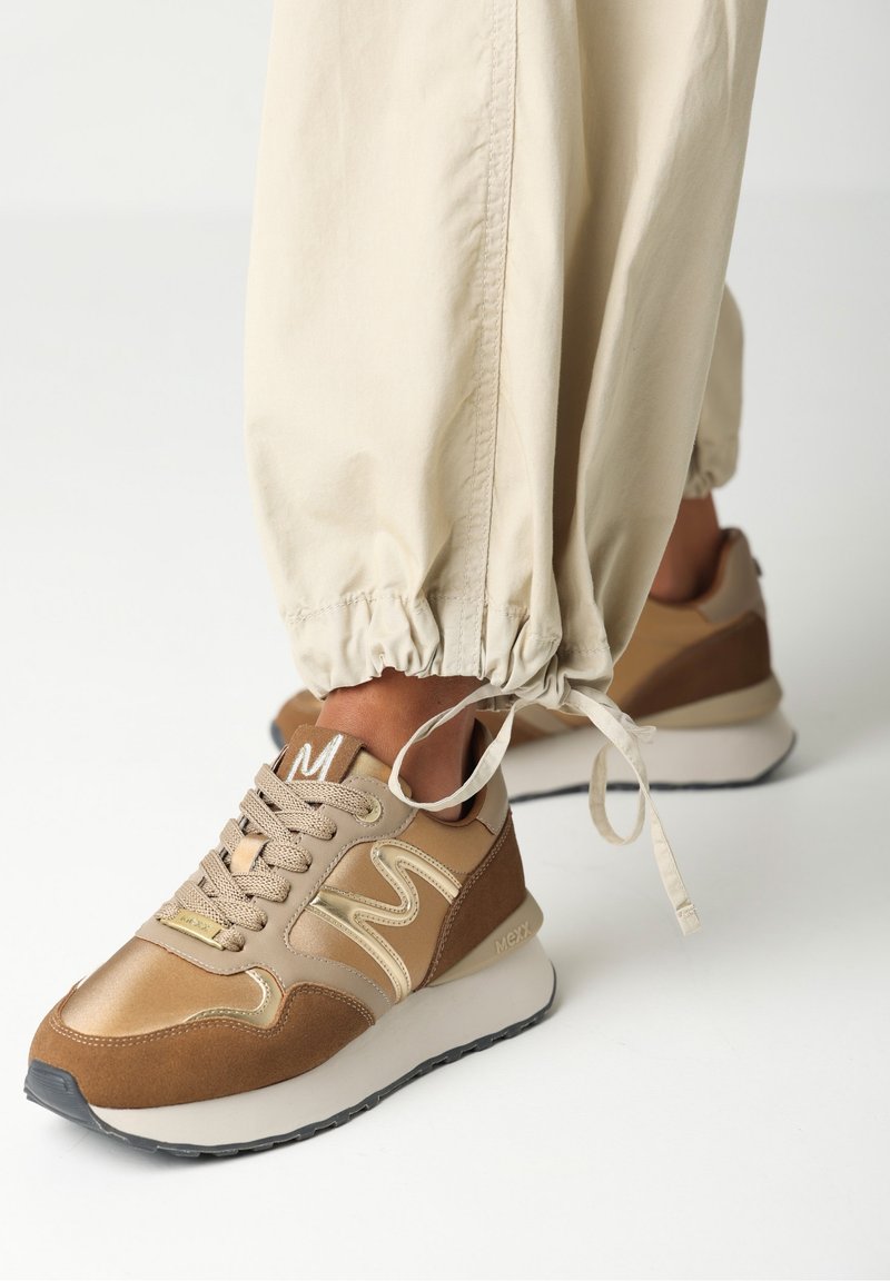 Baskets marron et beige avec semelles blanches épaisses portées avec un pantalon beige à cordon de serrage sur un fond uni.