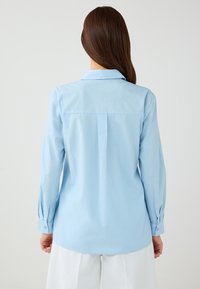 Woman with long brown hair wearing a light blue long-sleeve shirt and white pants, shown from the back against a plain background.