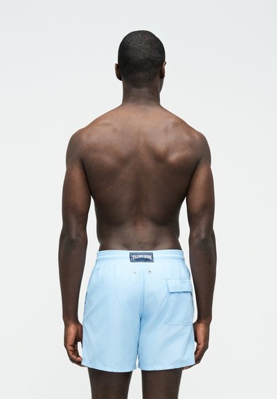 Άνδρας φαίνεται από πίσω φοράει ανοιχτό μπλε μαγιό με πίσω τσέπη και λογότυπο Vilebrequin στη ελαστική μέση.