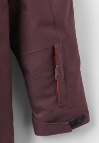 Donker bordeaux stijl jacket met diagonale weeftextuur, voorzien van een klein ritsvakje met rode voering en een zwarte klittenbandstrap bij de manchetten.